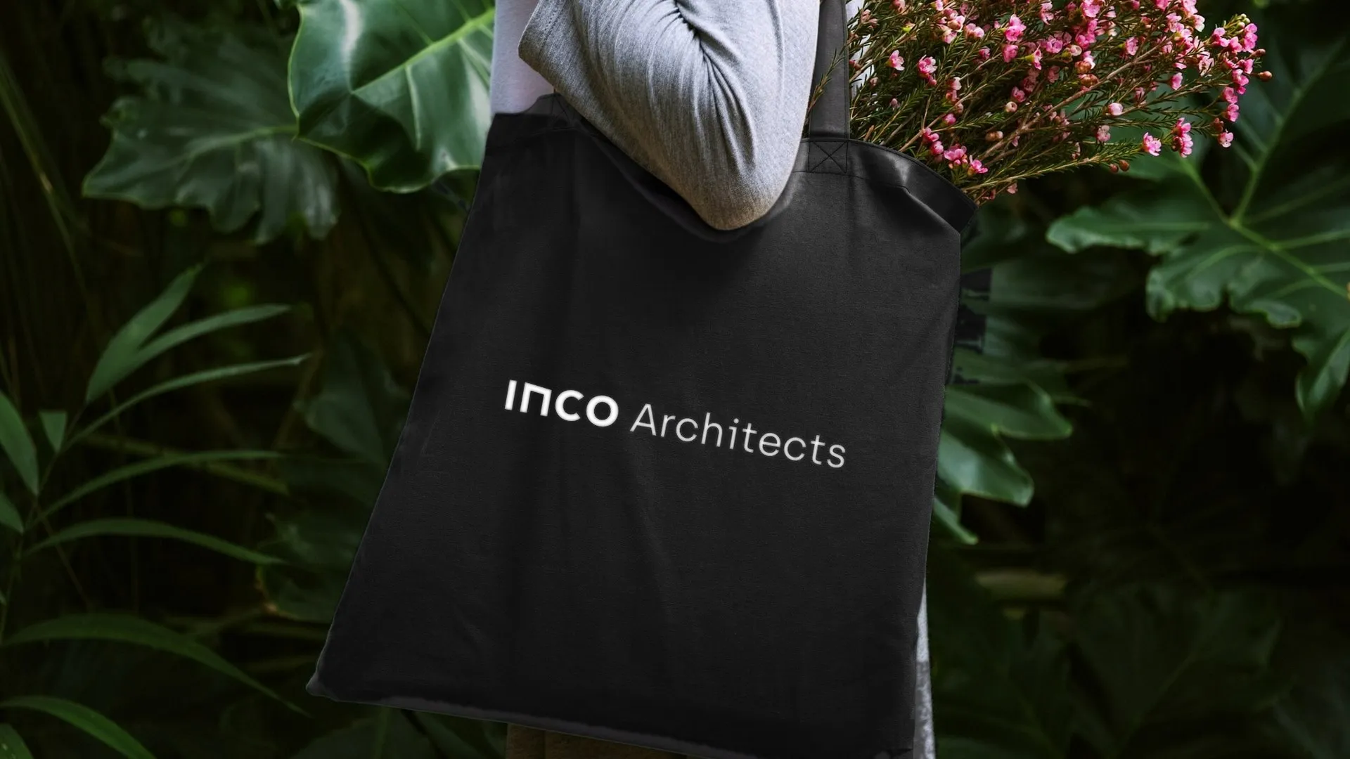 Czarna bawełniana torba z białym logo Inco Architects niesiona wśród zielonych roślin
